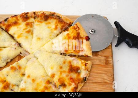 Une vue rapprochée montre un coupe-pizza tranchant une pizza au fromage sur une planche de bois. Banque D'Images