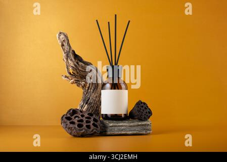 Diffuseur d'arôme en bouteille en verre brun avec des bâtons, bois flotté et gousses de lotus séchées sur fond orange, nature morte minimaliste avec des textures naturelles et Banque D'Images