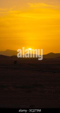 Coucher de soleil dans le désert du Sahara marocain avec des silhouettes de dunes de sable, des formes de montagne et un ciel doré brillant projetant une lumière chaude à travers l'horizon Banque D'Images