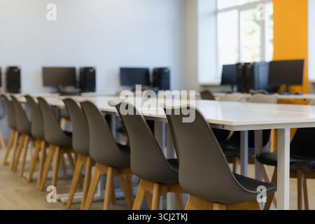Un espace de travail spacieux dispose de chaises élégantes et d'ordinateurs disposés soigneusement, éclairés par la lumière naturelle diffusant à travers de grandes fenêtres. Banque D'Images