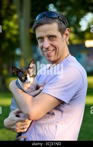 Un homme aux cheveux courts portant une chemise violet clair tient son chat Sphynx canadien dans un portrait vertical à l'extérieur. La photo met en évidence la compagnie, Banque D'Images