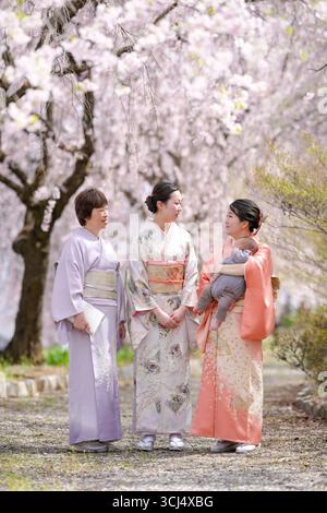 Trois femmes vêtues de vêtements traditionnels japonais se tiennent devant des cerisiers en fleurs. L'une des femmes tient un bébé. La scène est paisible et s. Banque D'Images