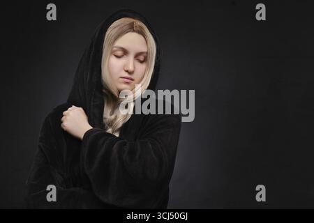 Belle jeune femme blonde dans la capuche noire regardant vers le bas sur fond noir avec espace de copie Banque D'Images