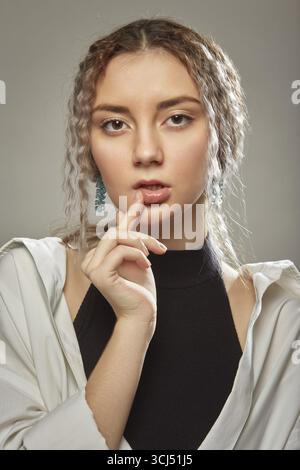 Jeune femme triste sérieuse avec les cheveux bouclés regardant la caméra avec le doigt sur la bouche Banque D'Images