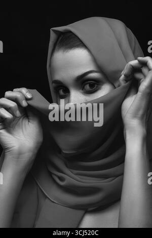 Belle jeune femme avec foulard gris couvrir son visage avec un maquillage de luxe sur fond noir regardant la caméra, monochrome Banque D'Images