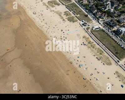 Vue aérienne d'une plage baignée de soleil grouillant de vie, où le sable doré rencontre la mer Azur, ombragée par la ville pittoresque de Bernières-sur-mer, Normandie, France. Banque D'Images