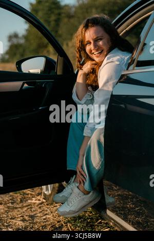 Femme souriante assise dans la porte de voiture à l'extérieur Banque D'Images