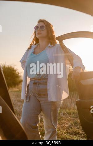 Femme en lunettes de soleil debout près de la voiture au coucher du soleil Banque D'Images