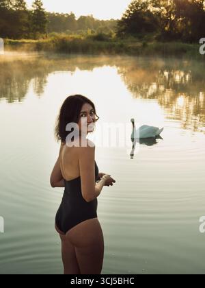 Femme souriante en maillot de bain noir et cygne sur le soleil Misty Lake à Dawn Banque D'Images