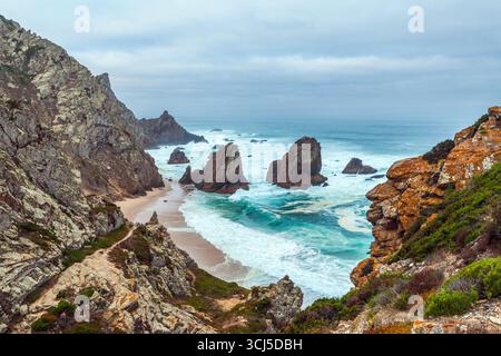 Une scène côtière magnifique avec des falaises accidentées, des vagues turquoises se écrasant sur une plage isolée Praia da Ursa, et des piles de mer spectaculaires sous un ciel doux Banque D'Images