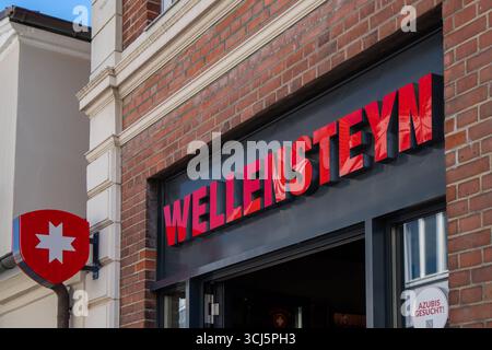 Travemünde, Deutschland 20 août 2025 : Im Bild : Leuchtendes Firmenschild des Modegeschäfts Wellensteyn in Travemünde. Deutlich sichtbarer roter Schriftzug WELLENSTEYN an der Backsteinfassade über dem Eingang, daneben das charakteristische rote Wappenlogo mit weißem Stern. DAS Geschäft befindet sich in zentraler Lage im Stadtteil Travemünde und bietet Outdoor- und Freizeitmode. IM Schaufenster ist zusätzlich ein Hinweis Azubis gesucht angebracht. Schleswig-Holstein *** Travemünde, Allemagne 20 août 2025 dans l'image signe lumineux de la société Wellensteyn magasin de mode à Travemünde clairement visible Banque D'Images