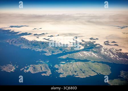 Côte Finnmark Norvège vue aérienne des montagnes et des fjords Oppeid / OPPEID, Norvège — une vue aérienne capture la côte accidentée du Finnmark, Norvège, regardant vers le nord en direction de la mer de Barents. Le premier plan présente les îles fragmentées de l'archipel de Loppa, caractérisées par des pentes vertes abruptes et des eaux turquoises peu profondes. Dominent le milieu du cadre sont les montagnes enneigées et les fjords profonds de l'île de Seiland et des péninsules adjacentes, Seiland étant connu pour ses petits glaciers. Un large pont de nuages recouvre la mer de Barents en arrière-plan, avec des sommets montagneux émergeant comme des îles. Le l Banque D'Images