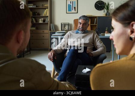 Homme indien d'âge moyen assis dans la chaise écoutant attentivement le jeune couple adulte caucasien pendant la session de conseil, tenant presse-papiers et stylo, étagère en arrière-plan Banque D'Images