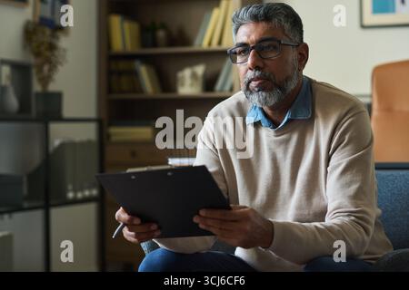 Homme indien d'âge moyen assis à l'intérieur tenant une planche à pince et un stylo, regardant attentivement vers l'avant, portant des lunettes et la barbe, semblant concentré pendant la réunion ou la consultation Banque D'Images