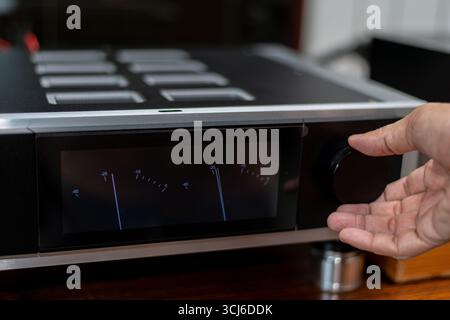 Gros plan d'une main tournant le bouton de volume sur un amplificateur hi-fi moderne avec vu-mètres analogiques, montrant la précision et la passion pour le son. Banque D'Images