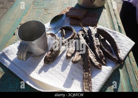Les gousses de caroube et les biscuits de caroube reposent sur une serviette en lin blanc à côté d'une tasse en étain usée et d'une fourchette sur une table en bois rustique. Banque D'Images
