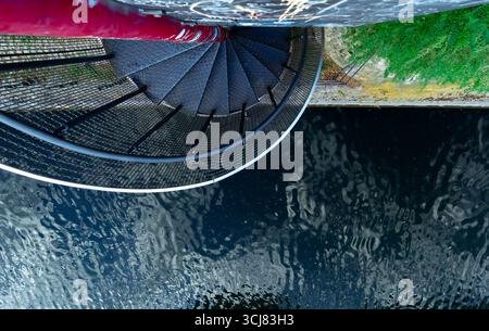 Vue en bas d'un escalier en colimaçon vers l'eau sombre ondulée avec des reflets et un bord urbain de rivière graveleux. Banque D'Images
