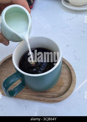 Gros plan du lait versé dans une tasse de café noir placée sur un plateau en bois, concept de nourriture et de boisson de style de vie. Banque D'Images