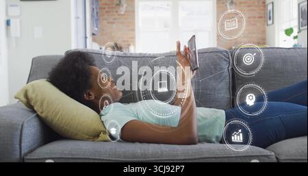 Adolescente couchée sur le canapé à la maison tenant le téléphone au-dessus de la tête avec oreiller jaune, icônes numériques Banque D'Images