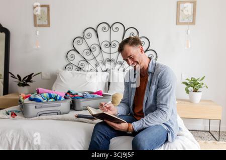 Homme assis sur le lit écriture dans un cahier avec valise ouverte dans la chambre, espace copie Banque D'Images