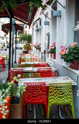 Terrasse de café colorée avec chaises en treillis rouge et vert bordant une rue européenne étroite ornée de boîtes à fleurs en géranium. Dynamisme urbain, café Banque D'Images