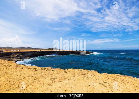 Côte rocheuse du parc national d'Arikok à Aruba près des ruines de moulin à or Bushiribana avec des voitures et bleu mer des Caraïbes. Banque D'Images