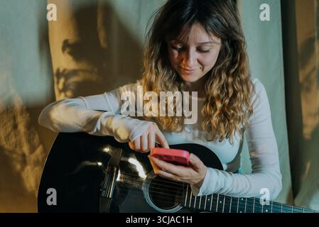 Jeune femme musicienne tenant une guitare acoustique et utilisant son smartphone, recherchant en ligne des accords de chanson ou des onglets tout en profitant de sa passion pour la musique dans un cadre détendu et décontracté Banque D'Images