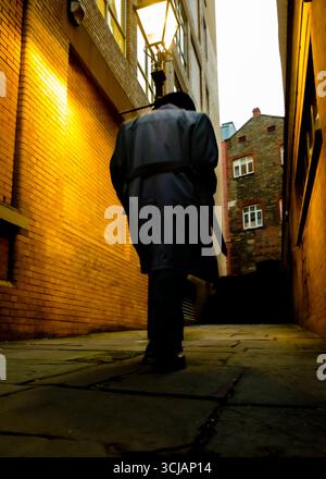 Une personne en long manteau marche dans une ruelle étroite, éclairée par un lampadaire. Banque D'Images