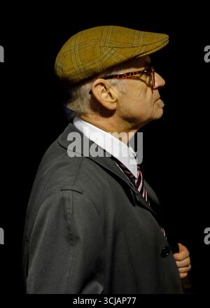 Un homme plus âgé portant une casquette en tweed, des lunettes et une veste de costume est vu de profil sur un fond noir. Banque D'Images