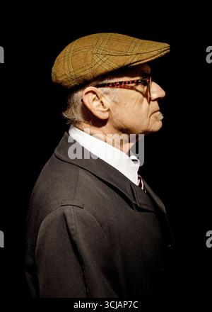 Portrait de profil de côté d'un homme âgé portant une casquette en tweed, des lunettes, une chemise à col blanc et un manteau foncé. Banque D'Images