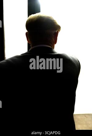 Un homme portant une casquette et un costume se tient dos à la caméra, regardant une lumière vive. Banque D'Images