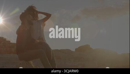 Embrassant couple portant des lunettes de soleil et des jeans sur le rocher du désert au coucher du soleil, avec des structures en pierre Banque D'Images