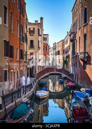 Une scène de canal classique à Venise, en Italie, avec une rangée de gondoles garées et des bâtiments historiques qui bordent le bord de l'eau. Le petit pont de pierre i. Banque D'Images