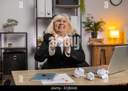 Femme d'affaires senior en colère travaillant au bureau à la maison jetant du papier froissé ayant une dépression nerveuse au travail migraine, gestion du stress, problèmes de détresse mentale, perdre son sang-froid, réaction à l'échec. Banque D'Images