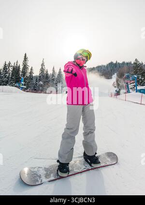 Un snowboarder se tient sur une pente enneigée, vêtu d'une veste rose éclatante et de lunettes, saisissant une pose ludique Banque D'Images
