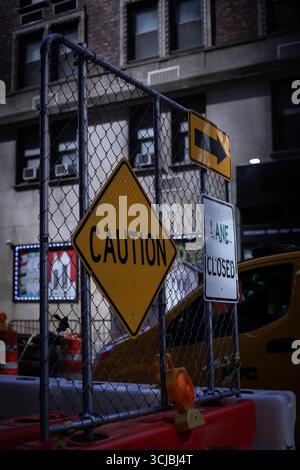 Un trio de panneaux routiers urbains – attention, Lane Closed et une flèche directionnelle – montés sur une clôture en maille créent une scène visuellement saisissante devant une ville b. Banque D'Images