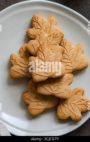 Vue de dessus de biscuits en forme de feuille d'érable disposés sur une plaque blanche. Les cookies sont beiges avec des empreintes de feuilles détaillées, reposant sur un fond awooden. Banque D'Images