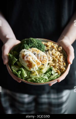 Mains coupées méconnaissables tenant un bol avec du fromage halloumi grillé, du brocoli, du quinoa et des verts feuillus, sur un fond sombre, représentant Banque D'Images