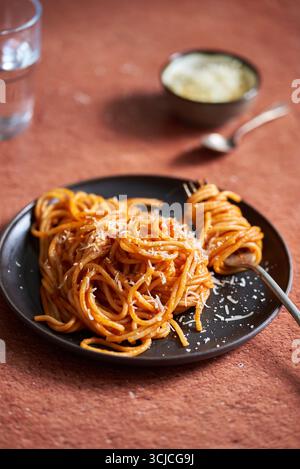 Une portion de spaghetti garnie d'une riche sauce tomate et de parmesan râpé élégamment présenté sur une assiette sombre. L'arrière-plan comporte un chaud, t Banque D'Images