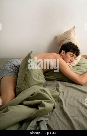 Un jeune homme repose paisiblement sur un lit confortable, entouré d'oreillers moelleux et de draps confortables. La lumière du matin sereine illumine doucement la pièce, c Banque D'Images