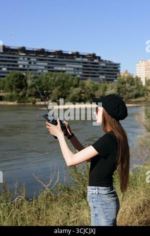 Une jeune femme se tient au bord de la rivière, actionnant intentionnellement un dispositif de radiocommande. C'est une journée d'été ensoleillée, sa tenue décontractée reflétant la détente à Banque D'Images