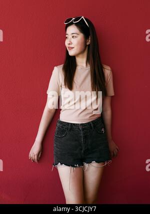 Une jeune femme élégante se tient en toute confiance contre un mur rouge frappant, sa tenue décontractée complétant parfaitement la toile de fond vibrante, exsudant un sens Banque D'Images