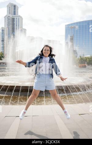 Touriste joyeuse sautant de joie devant une fontaine à Ekaterinbourg, en Russie. Entouré de bâtiments modernes et la lumière du soleil, parfait pour tra Banque D'Images