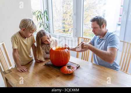Une famille profite d'une activité automnale amusante en sculptant une grande citrouille orange pour Halloween. Le père et les enfants s'engagent dans la créativité, ce qui rend durable moi Banque D'Images