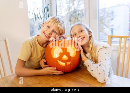 Deux enfants souriants aiment sculpter une grande citrouille joyeuse ensemble à la maison pour Halloween. La citrouille décorée ajoute une touche festive à l'autu confortable Banque D'Images