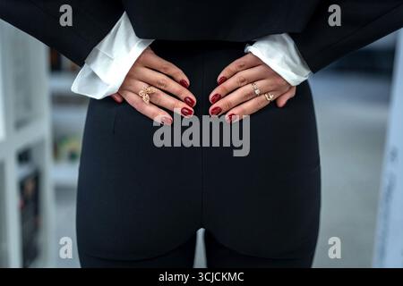 Une femme élégante dans un costume noir est vue à l'intérieur, ses mains reposant sur son dos, mettant en valeur ses bijoux élégants et vernis à ongles rouge vif, exsudant co Banque D'Images