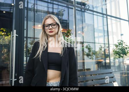 Jeune femme blonde dans une tenue noire élégante et des lunettes debout en toute confiance à l'extérieur. Elle pose près d'un bâtiment de verre moderne par une journée ensoleillée, showcasin Banque D'Images