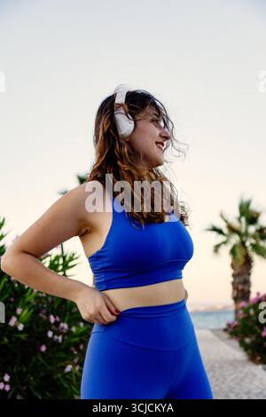 Une jeune femme en sportwear bleu profitant d'une séance d'entraînement en plein air au bord de la plage au coucher du soleil. Elle porte des écouteurs, embrasse la forme physique et un mode de vie sain Banque D'Images