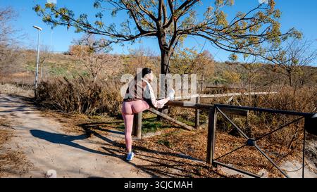 Une athlète étire sa jambe sur une clôture en bois dans un parc pittoresque, se préparant pour une course. Le ciel bleu clair et les feuilles d'automne créent une revigoratine Banque D'Images