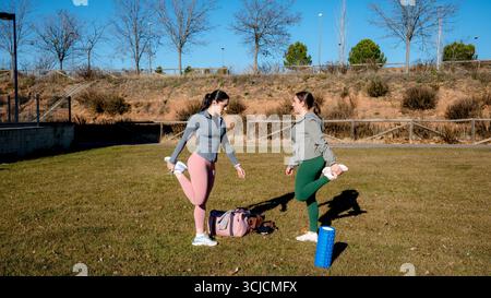 Deux femmes s'étirent dans un parc ensoleillé avant de courir. Les deux portent des vêtements de sport et tiennent un pied derrière eux, en se concentrant sur la flexibilité. Un sac de gym et un rouleau de mousse Banque D'Images
